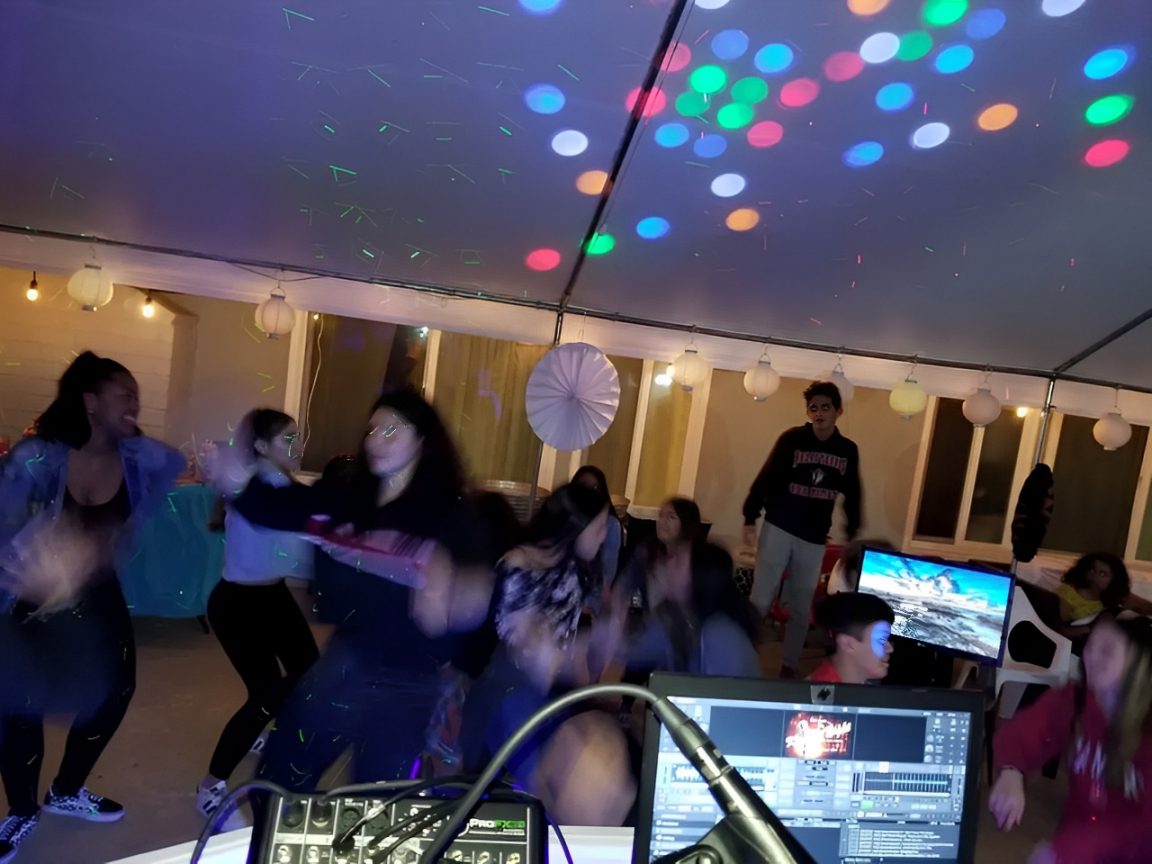 People are dancing under a tent with colorful lights and a DJ setup visible in the foreground.