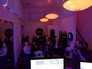 People are gathered in a dimly lit room with colorful lights. Large lamps hang from the ceiling, and a computer setup with monitors is visible in the foreground.