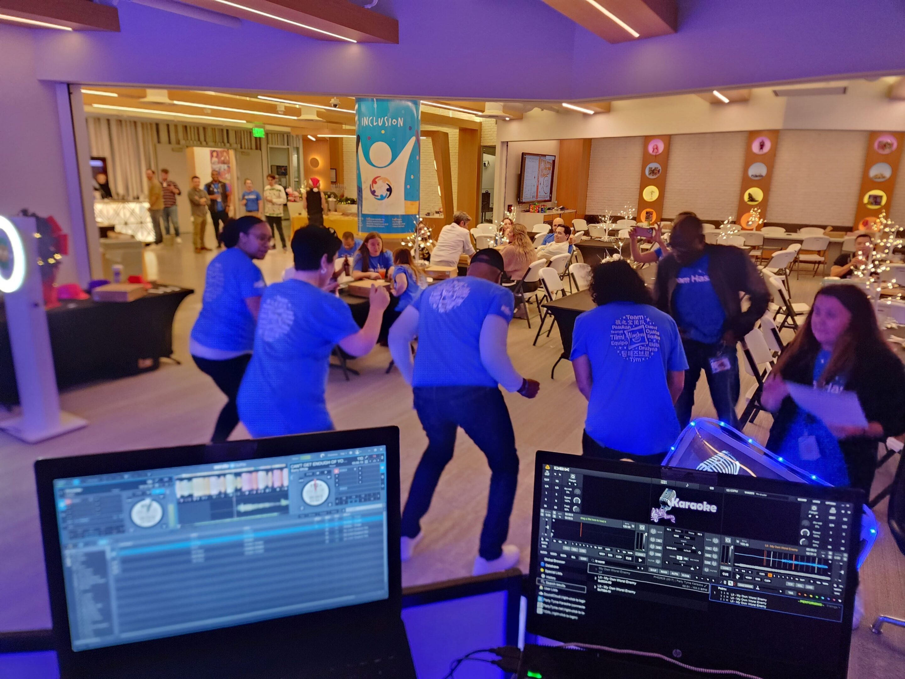 People dance in a brightly lit room during an indoor event, viewed from behind a DJ station music equipment and the song "wobble"