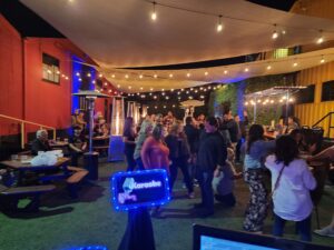 People gather outdoors under string lights and canopy for a nighttime karaoke event; some are seated at picnic tables, others stand and socialize.