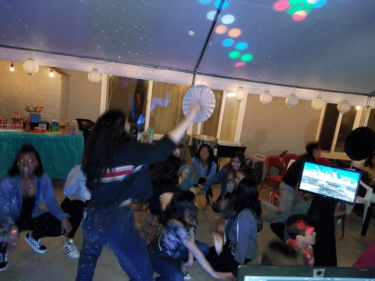 A group of people, mostly children, dance under a tent with string lights and colorful disco lights at a party. A decorated table and large paper fans are visible in the background.