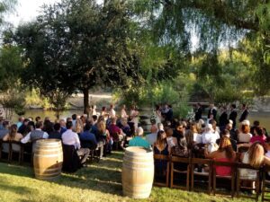 Outdoor wedding ceremony by a river with guests seated on wooden chairs, a wedding party at the front, and wine barrels in the foreground under trees.