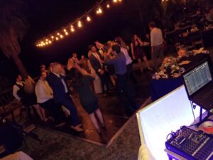 A group of people dance on an outdoor dance floor at night under string lights, with a DJ setup and sound equipment visible in the foreground.