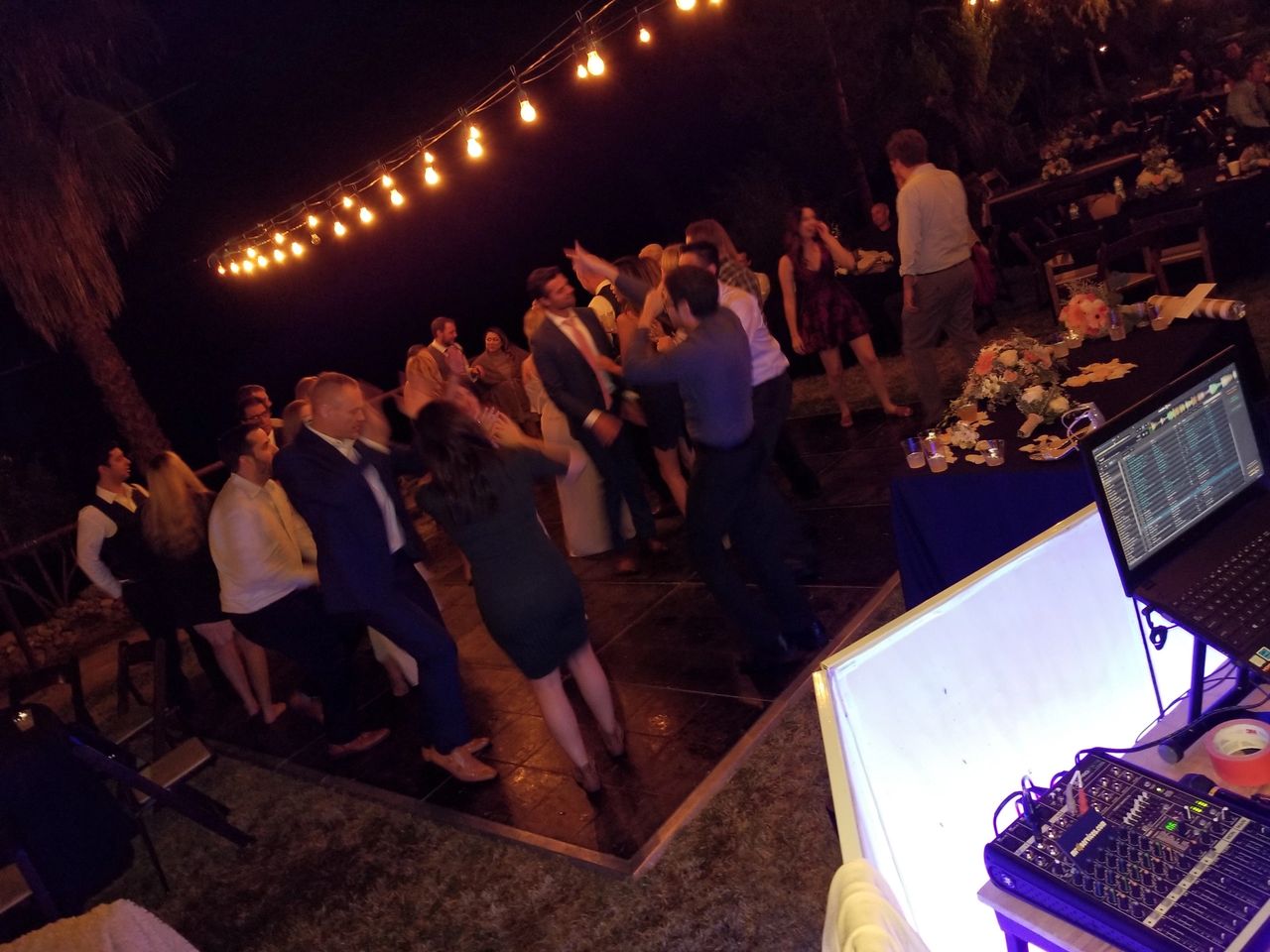 A group of people dance on an outdoor dance floor at night under string lights, with a DJ setup and sound equipment visible in the foreground.