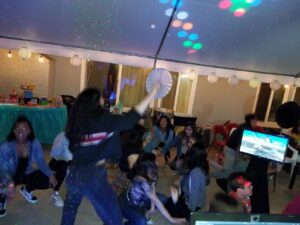 A group of people dance under colorful lights at an indoor party, with decorations and a table of snacks in the background.
