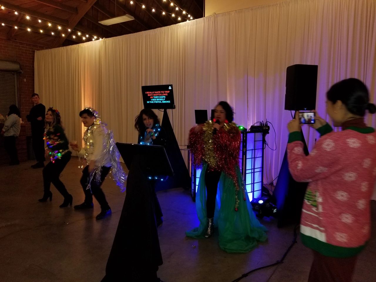 Four people in colorful, shiny costumes perform karaoke on a stage, while a woman in a festive sweater takes a photo with her phone. White drapes and string lights decorate the venue.