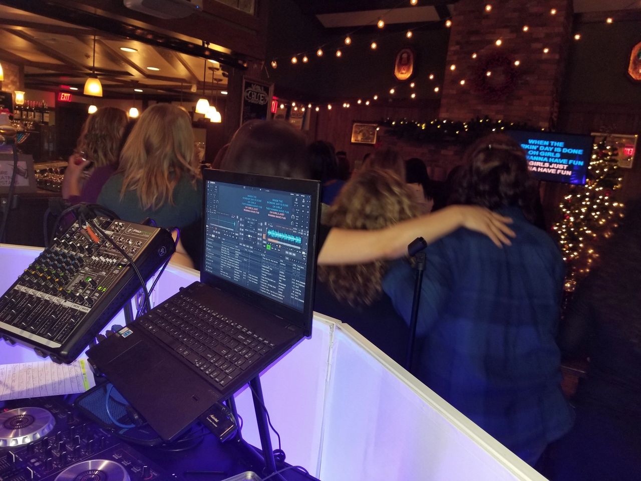 A group of people with arms around each other sing karaoke at a bar, viewed from behind a DJ booth with audio equipment and a laptop.