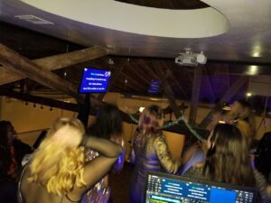 A group of people sing karaoke in a dimly lit room, facing a screen displaying song lyrics. A projector and DJ setup are visible in the background.