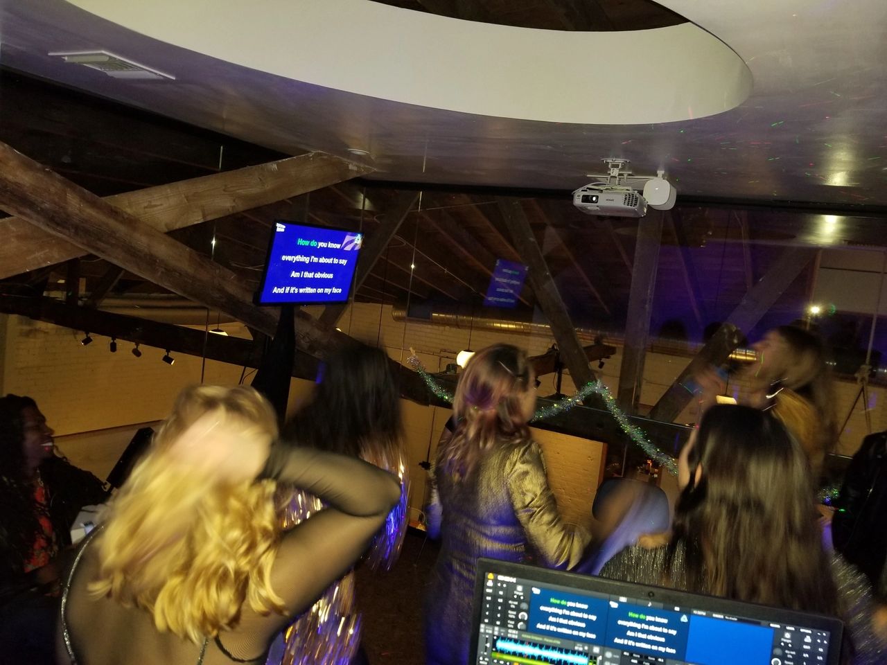 A group of people sing karaoke in a dimly lit room, facing a screen with song lyrics projected, under exposed wooden beams and a circular ceiling feature.