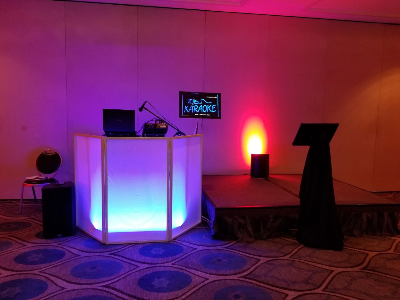 A karaoke setup with a lit DJ booth, microphone, laptop, monitor displaying “KARAOKE”, and a small stage with a music stand in a room with patterned carpet.