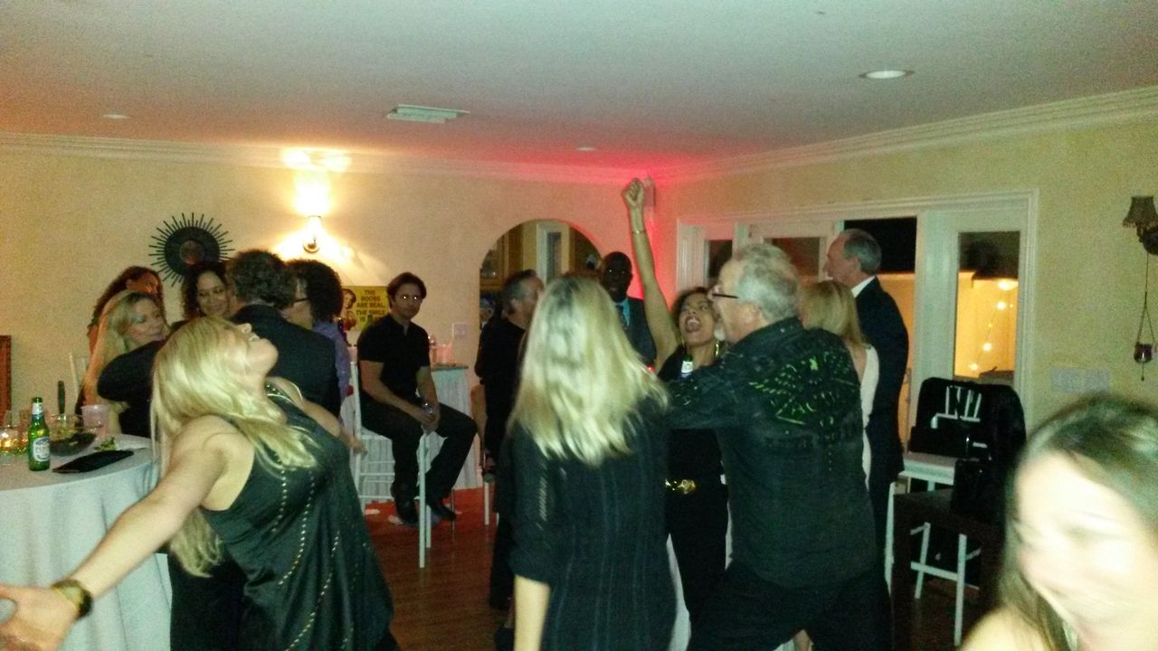 A group of people dance and socialize in a dimly lit room at a party; drinks are visible on a nearby table.