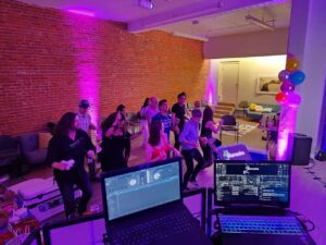 A group of people dance in a room with exposed brick walls and purple lighting, viewed from behind DJ equipment. Balloons and snacks are visible in the background.