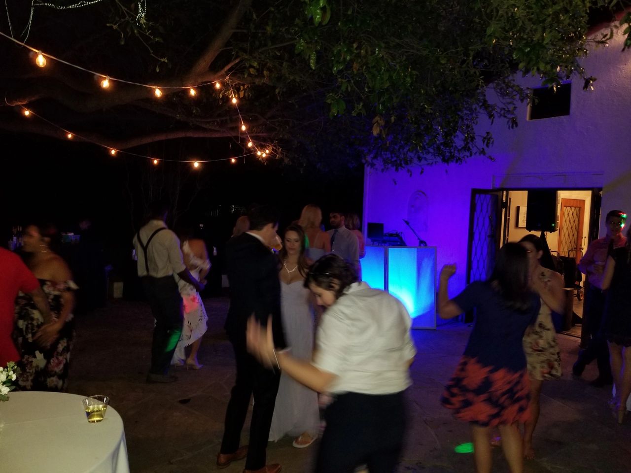 People dance outdoors at night under string lights, with a lit DJ booth and a white building in the background. Drinks and tables are visible in the scene.