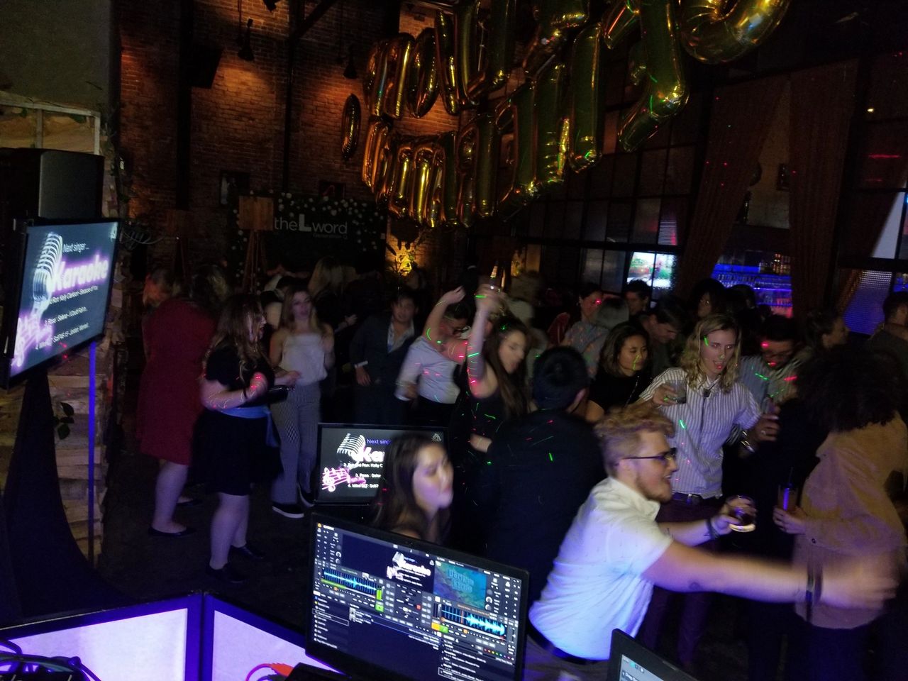 A crowd of people dance and socialize at a dimly lit indoor party with gold balloons and a karaoke setup visible in the foreground.