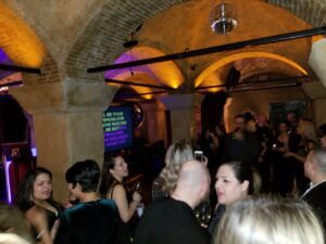 A group of people socializes at a dimly lit indoor party with arched ceilings and a disco ball; a karaoke screen displays song lyrics in the background.