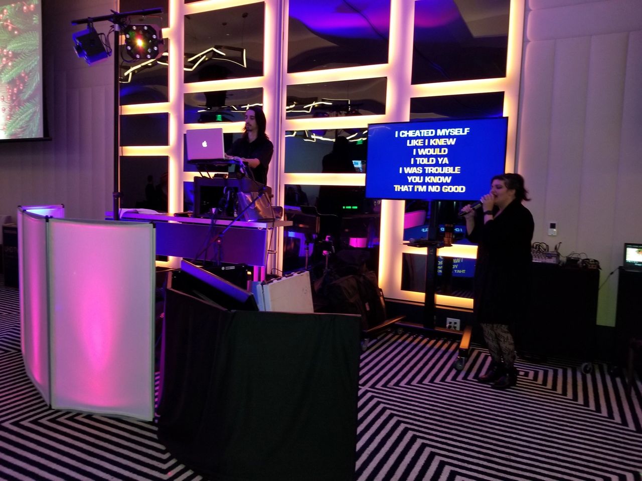 A woman sings into a microphone while another operates a laptop and DJ equipment; karaoke lyrics are displayed on a large screen behind them.
