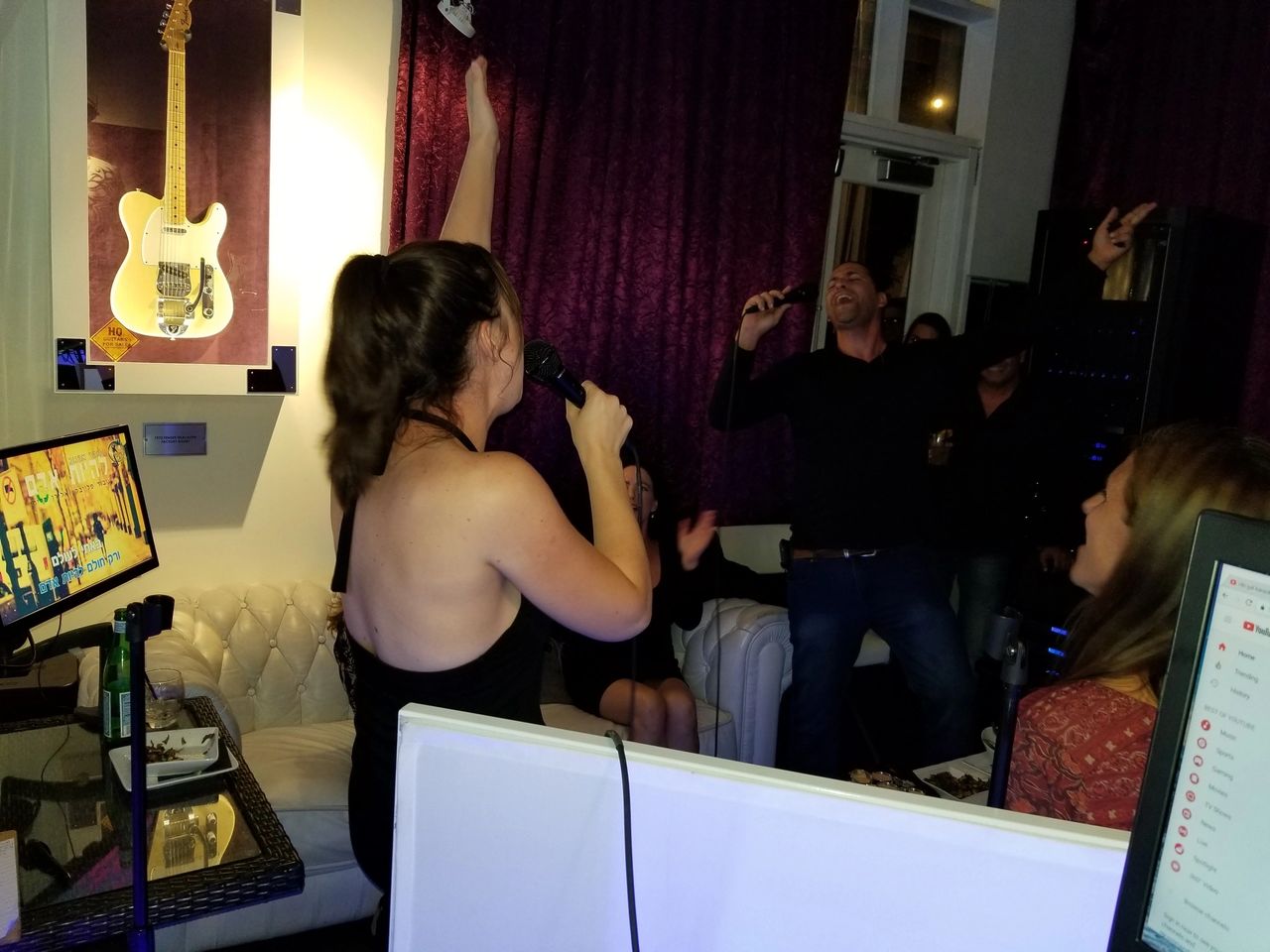 People singing and cheering in a karaoke room with microphones, a guitar displayed on the wall, and screens showing lyrics.