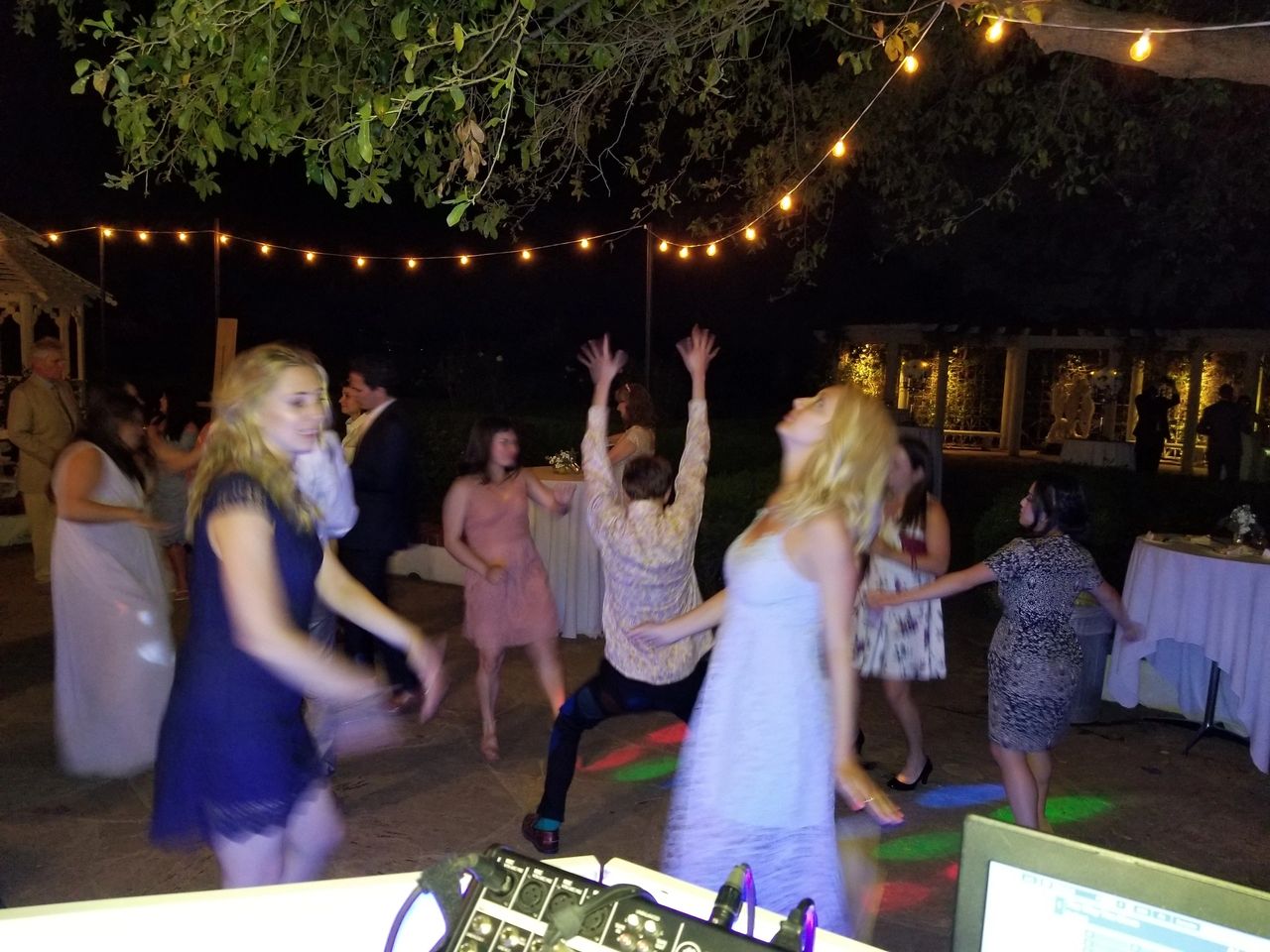 A group of people dance energetically on an outdoor patio at night, surrounded by string lights, with tables and trees in the background.