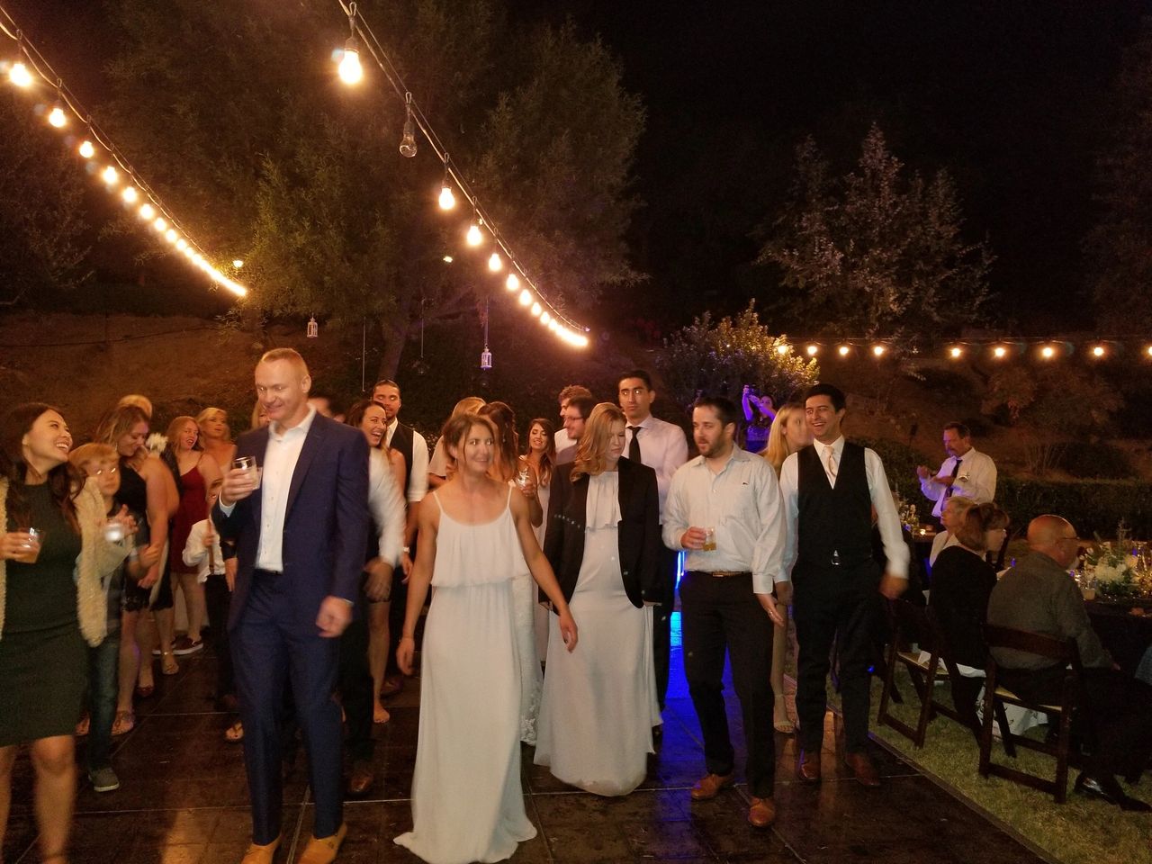 A group of people in formal attire walk together on a lit outdoor floor at night, with string lights overhead and trees in the background.