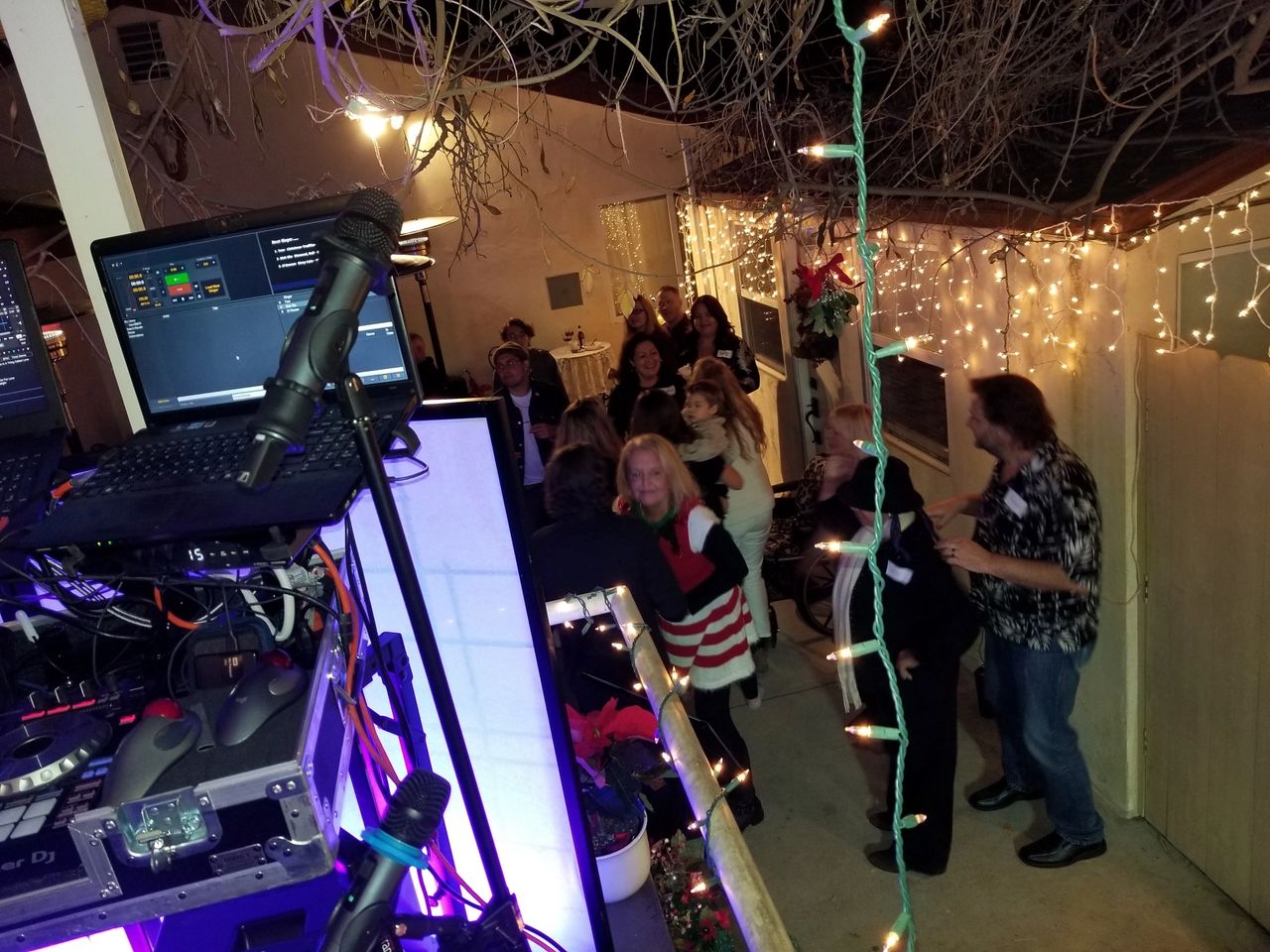 A group of people socialize and dance outdoors at night near a DJ booth, surrounded by string lights and festive decorations.