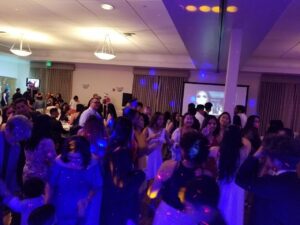 A large group of people are dancing and socializing in a banquet hall with colorful lights and a projection screen in the background.