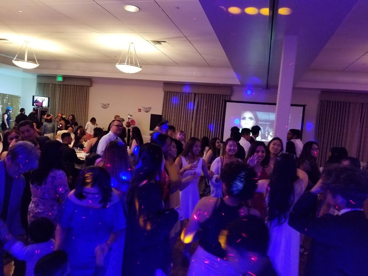A large group of people are dancing and socializing in a banquet hall with colorful lights and a projection screen in the background.