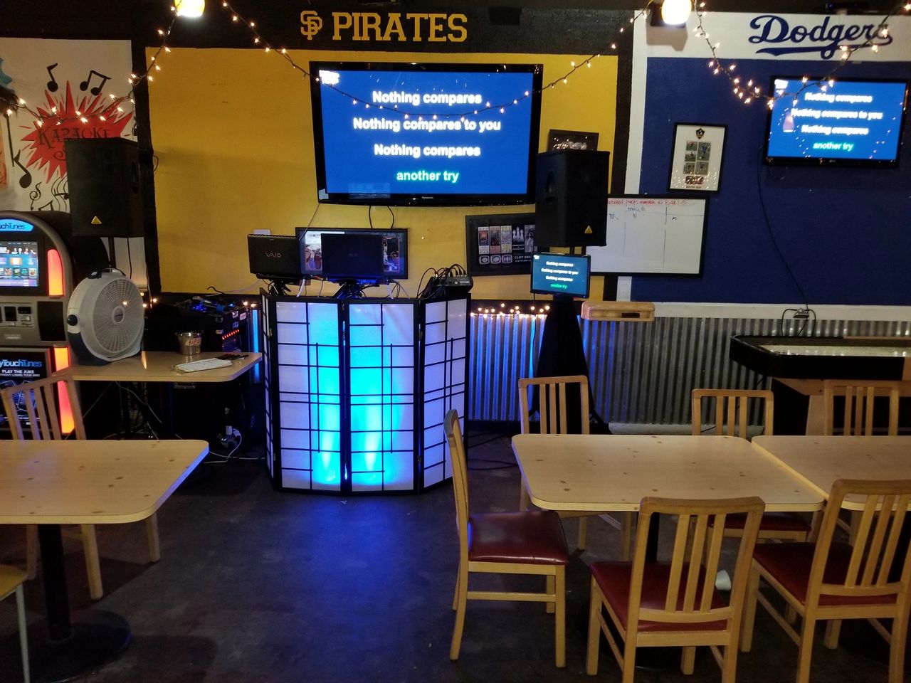 A karaoke setup in a casual dining area with lyrics on TV screens, wooden tables and chairs, and sports team signs on the walls.