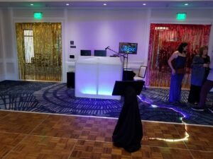 A karaoke setup with a lit DJ table, screen, and microphone stand on a dance floor; gold and red metallic streamers hang over two doorways; a few people stand nearby.