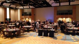 People gather and socialize around banquet tables in a large, carpeted event hall with a wooden dance floor in the foreground.