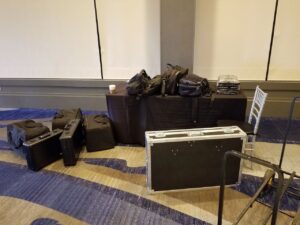 A table and floor hold several black equipment cases, bags, folded cloths, and a cup; a folding chair is nearby on a blue-patterned carpet.