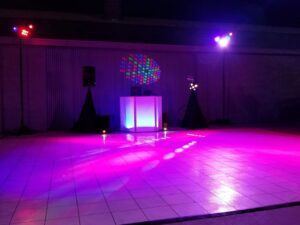 A DJ booth with speakers and colorful lights is set up on a tiled dance floor in a dimly lit indoor space.
