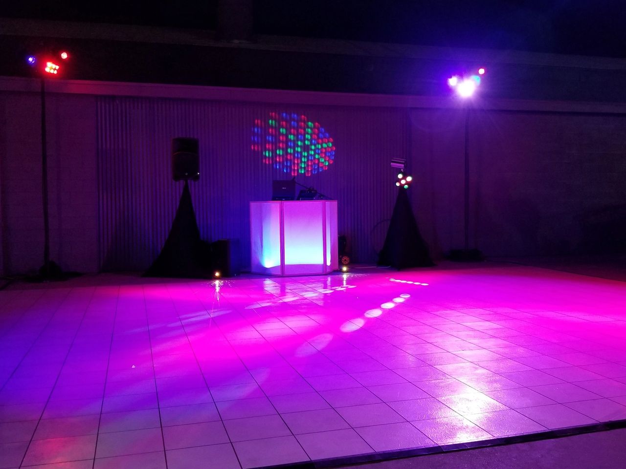 A DJ booth with speakers and colorful lights is set up on a tiled dance floor in a dimly lit indoor space.