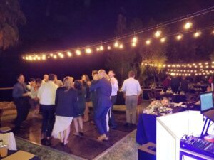 A group of people are dancing on an outdoor dance floor at night under string lights, with tables and a DJ booth nearby.