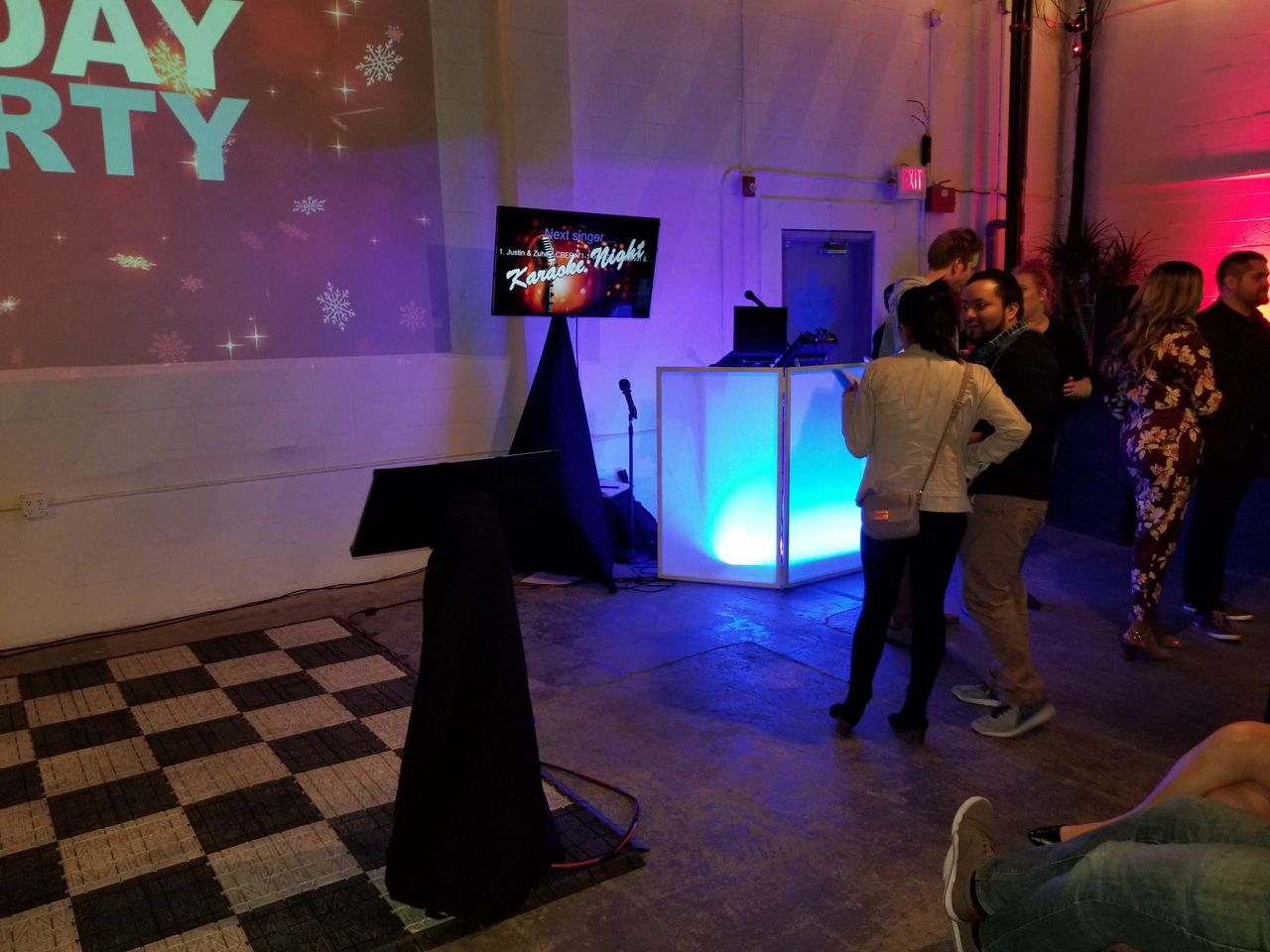 A group of people stands near a brightly lit DJ booth and karaoke setup in an indoor event space with party-themed decorations.