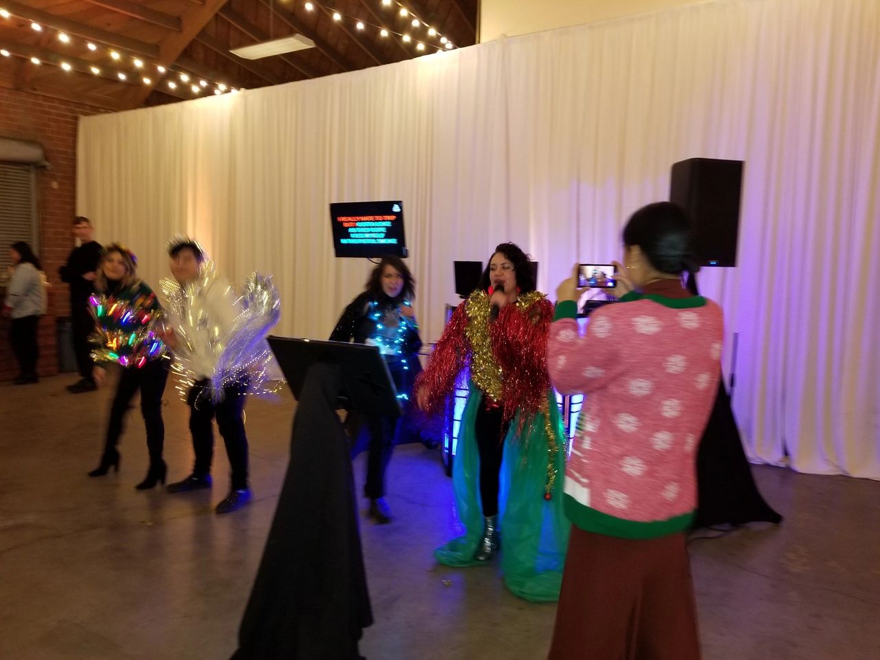Four people in festive costumes perform karaoke on a stage, while another person in a red sweater films them with a smartphone in a decorated indoor space.