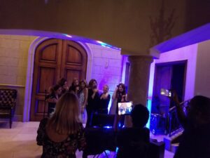 A group of women sing together at a karaoke setup in a room with stone walls and wooden doors, while others watch and record with their phones.