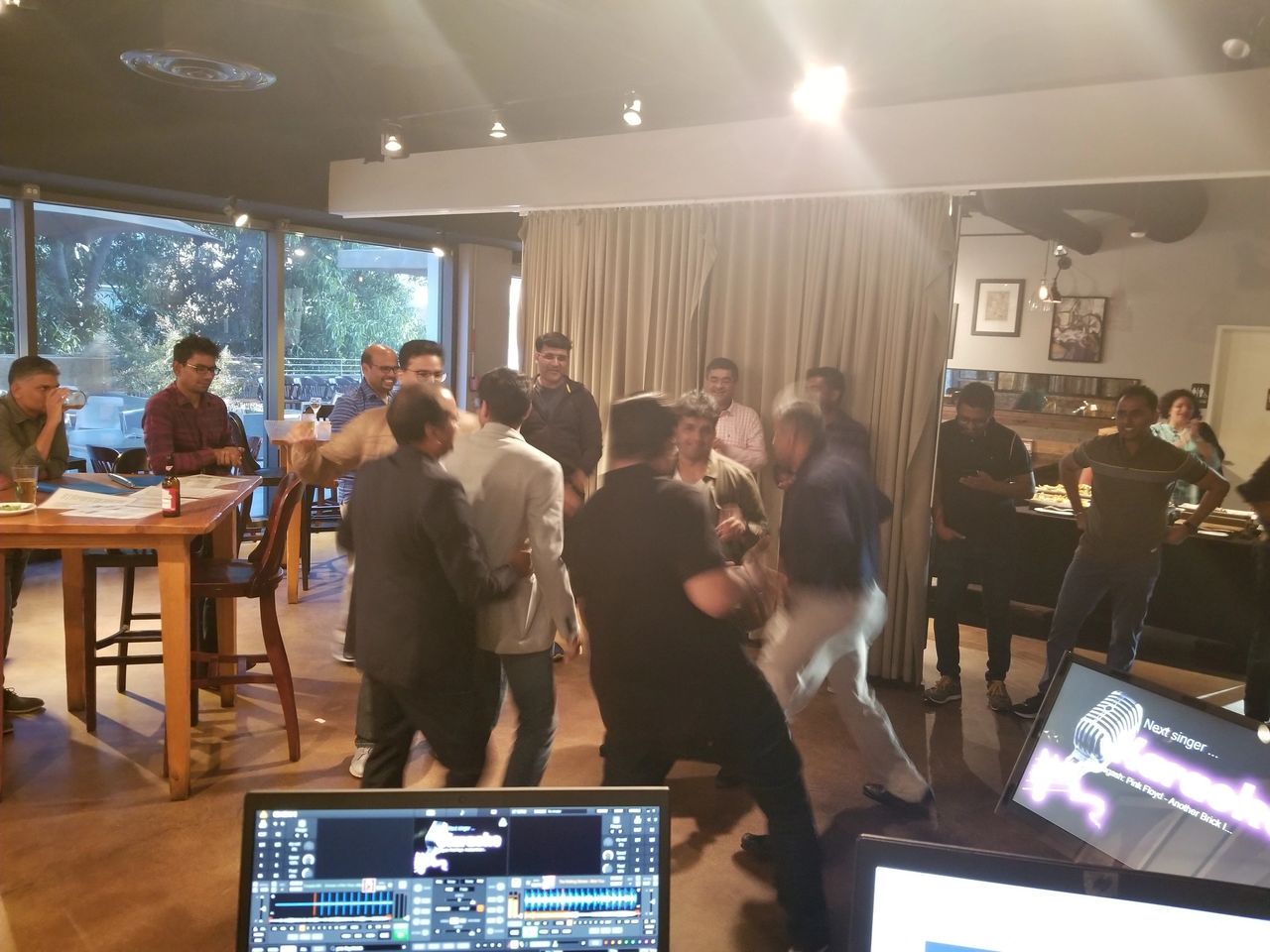 A group of people are gathered in a restaurant or cafe, some appearing to argue or confront each other while others watch. Tables, chairs, and computer monitors are visible in the foreground.