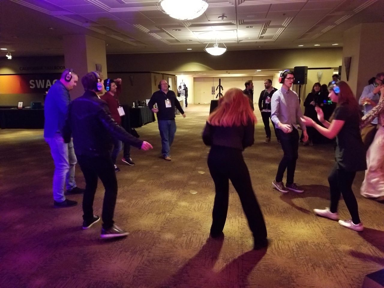 A group of people wearing headphones dance in a dimly lit room with brown carpet and overhead lights, participating in a silent disco event.