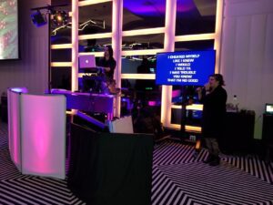 A woman sings karaoke while another operates a laptop at a DJ booth; song lyrics are displayed on a screen beside her in a room with colorful lighting.