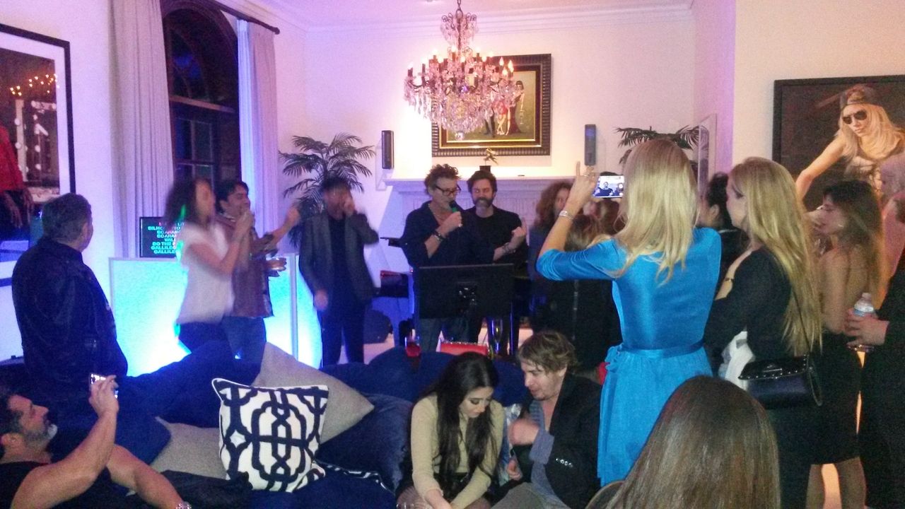 A group of people gather in a living room at night, some singing karaoke under a chandelier, while others watch, sit, or take photos.