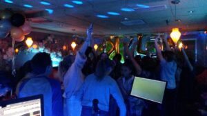 A group of people dance and raise their arms at a dimly lit party with balloons, decorations, and blue lighting.