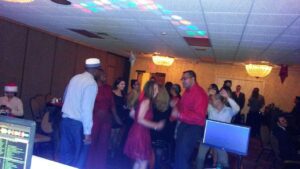 A group of people are dancing in a dimly lit event room with chandeliers, while others sit at tables. A DJ booth and computer screens are visible in the foreground.