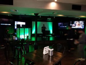 Dimly lit bar with empty tables and chairs, green-lit stage in the center, one person on stage, several TV screens on surrounding walls, and a few people moving around.