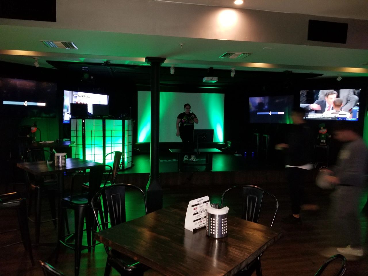 Dimly lit bar with empty tables and chairs, green-lit stage in the center, one person on stage, several TV screens on surrounding walls, and a few people moving around.