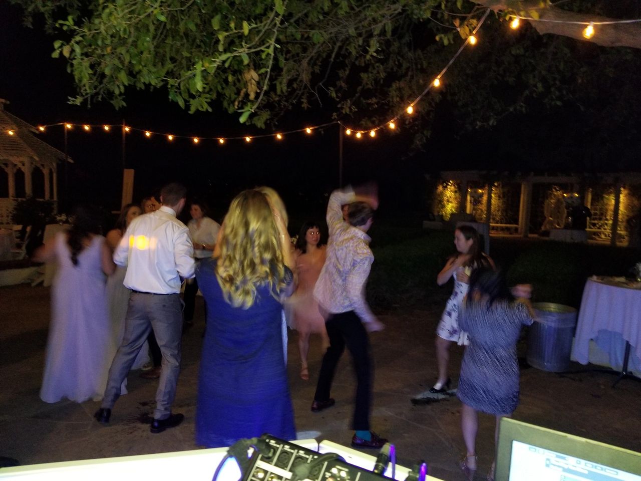 A group of people dance outdoors at night under string lights, with a DJ booth visible in the foreground and trees overhead.