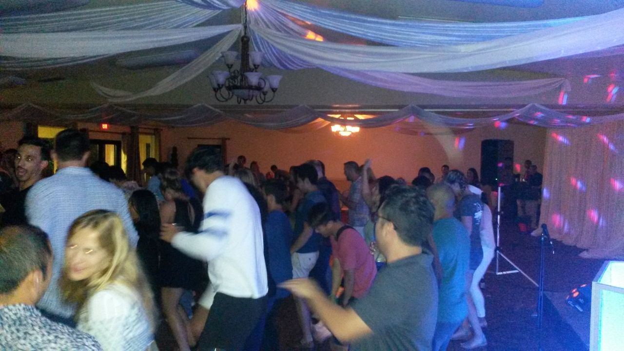 A large group of people are dancing in a decorated indoor venue with draped white fabric on the ceiling and colorful lighting effects.