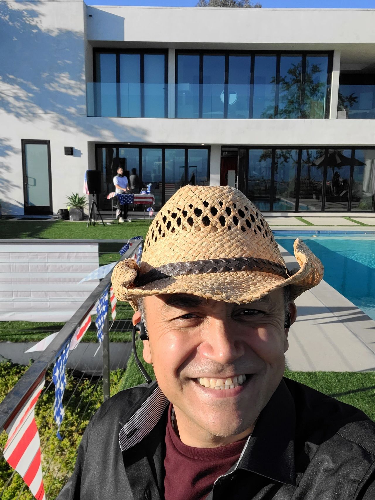A man in a straw hat smiles for a selfie by a pool, with a modern house, American flags, and a party DJ playing music near a table in the background.