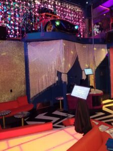 A colorful karaoke lounge with red sofas, a glowing dance floor, a stage area, and a car displayed on an upper ledge amid hanging decorations and shimmering curtains.