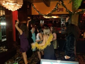 A group of women in dresses and costumes dance and raise their arms in a dimly lit, festively decorated room.