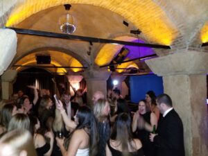 The Karaoke Dj Party for entrepreneurs. Guests dressed in formal attire are dancing and socializing at a lively party in a stone-arched room with yellow lighting and a disco ball.